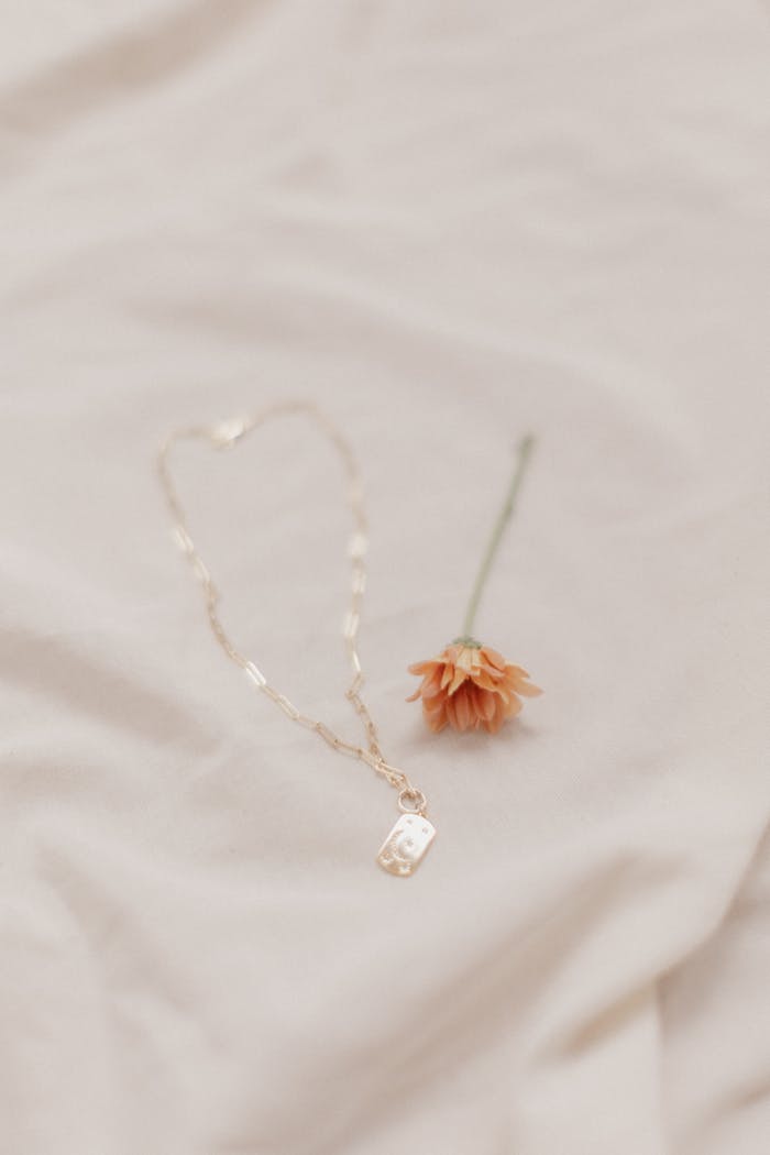 Close-up of gold necklace with pendant and orange flower on soft fabric, minimalist style.